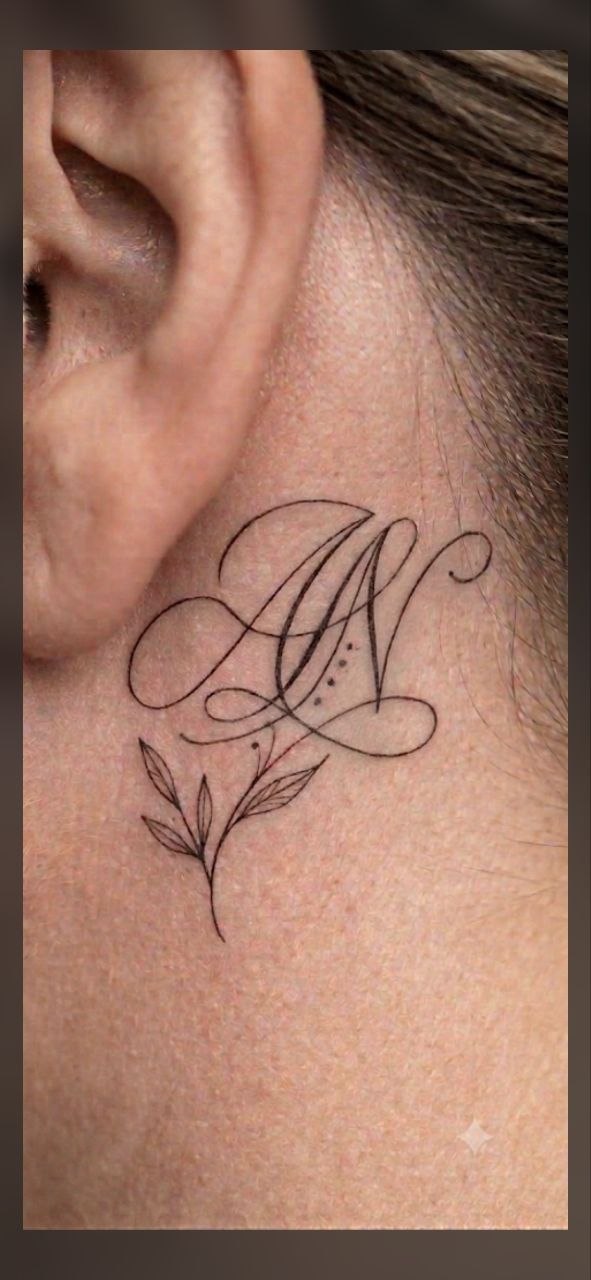 Fineline tattoo: sierlijke initialen met botanisch takje achter het oor